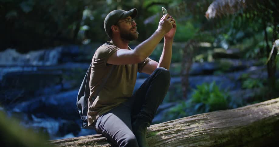 A man is sitting on a log near water taking pictures with his phone