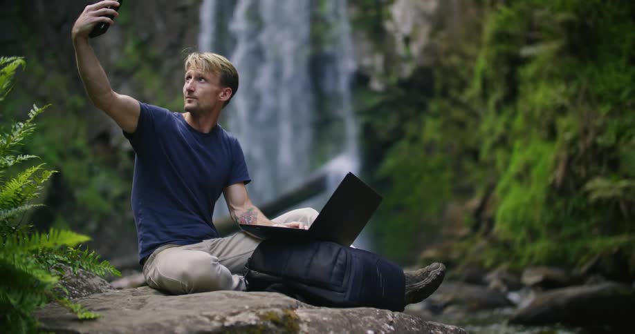 A man is sitting on a ledge near a waterfall typing on his laptop computer and holding his phone