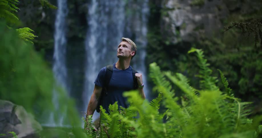 A man wearing a backpack is standing near a waterfall in the woods looking around