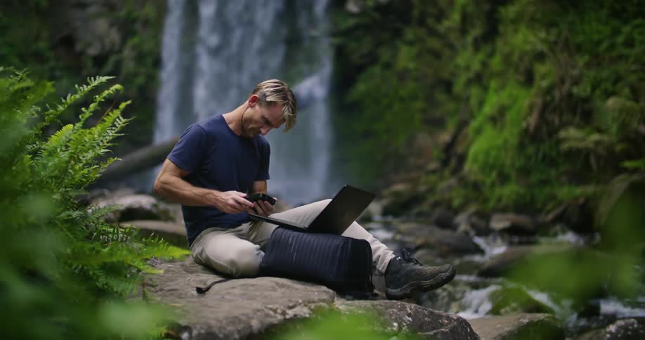 A man types on a laptop while sitting beside a stream near a waterfall during the day