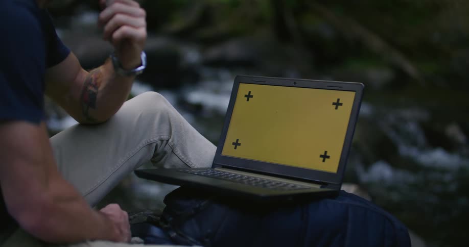 Man sits near stream with laptop propped on backpack