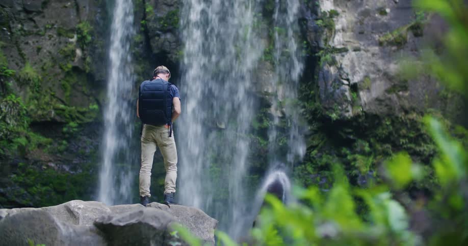 Blonde man wearing dark blue backpack t shirt and brown pants takes photo of waterfall with mobile phone in the forest