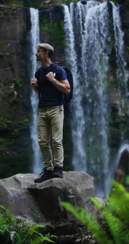 A man is perched on a large rock by the waterfall