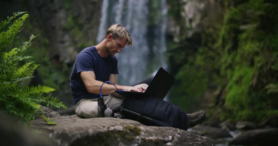 A man works on his laptop while sitting near a waterfall