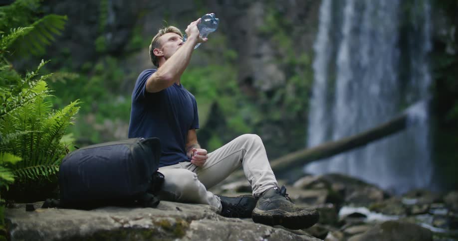 A man is sitting on a ledge near waterfalls and a stream as he takes a bottle of water from his backpack and has a drink