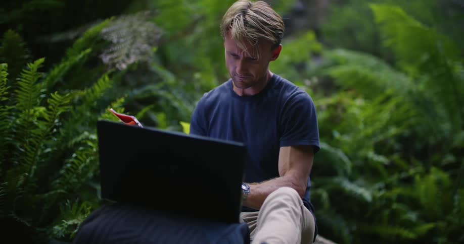 Man sits outdoors near ferns reading from file and typing on laptop