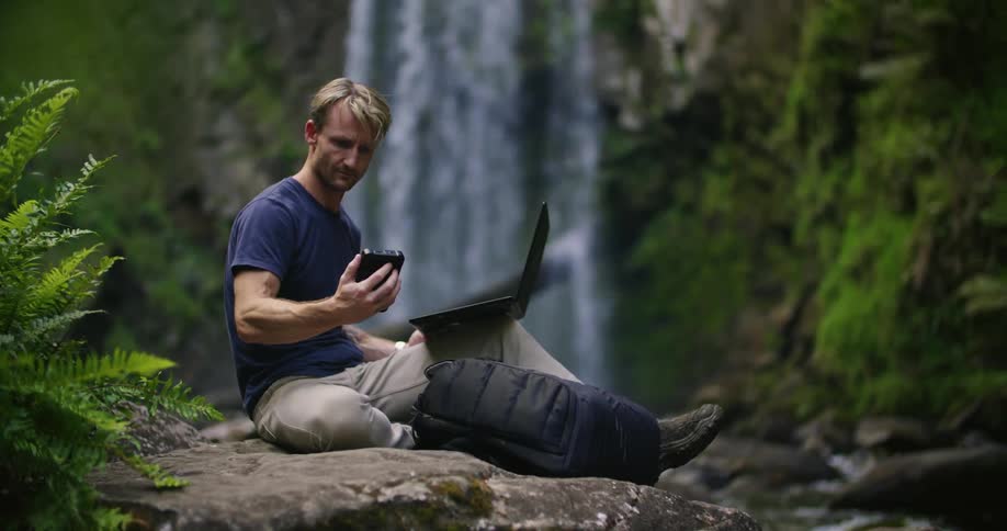 A man is typing on a laptop while sitting on a large boulder with a waterfall in the background