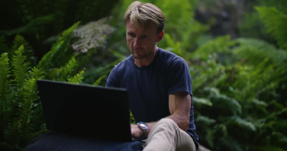 Blonde man sitting outdoors near ferns while typing on a laptop looks up briefly