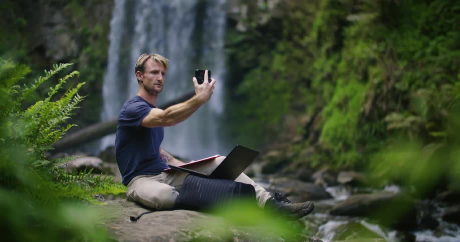 Man sitting on riverbank near waterfall checks area with device before typing on laptop