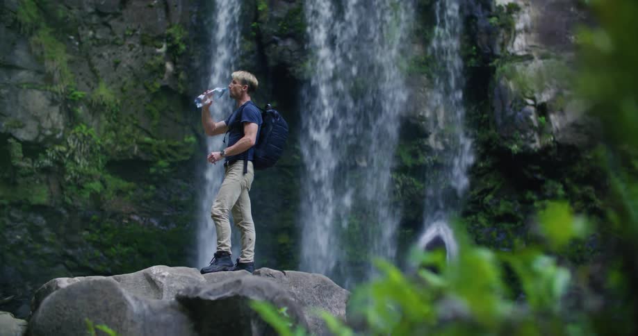 A man in hiking gear is standing on a large boulder and drinking water while looking up at the waterfall above him