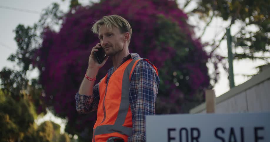 Man wearing safety vest talks on smartphone nearfor sale sign for bank foreclosure property
