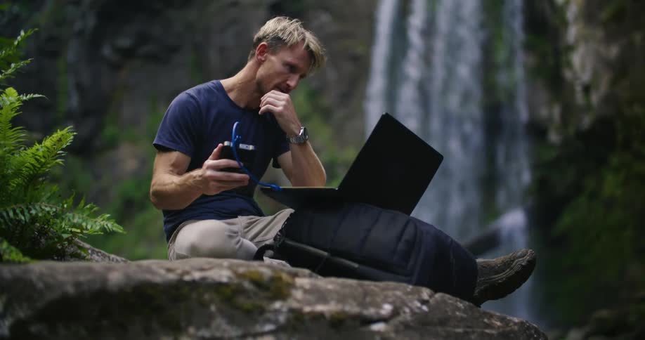 A hiker is sitting on a large boulder and typing on a laptop with a tall waterfall in the background
