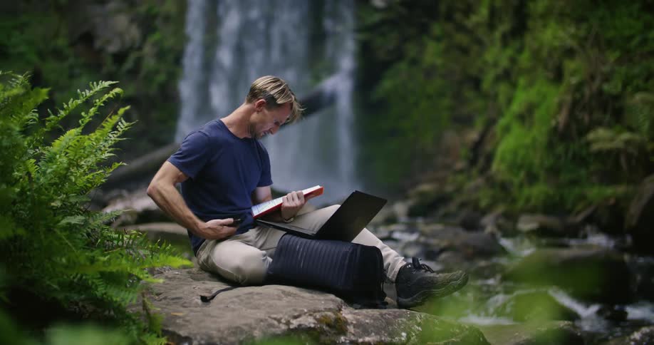 A man is sitting on a ledge near a waterfall and stream holding a notebook as he types on his laptop computer and holds up his phone