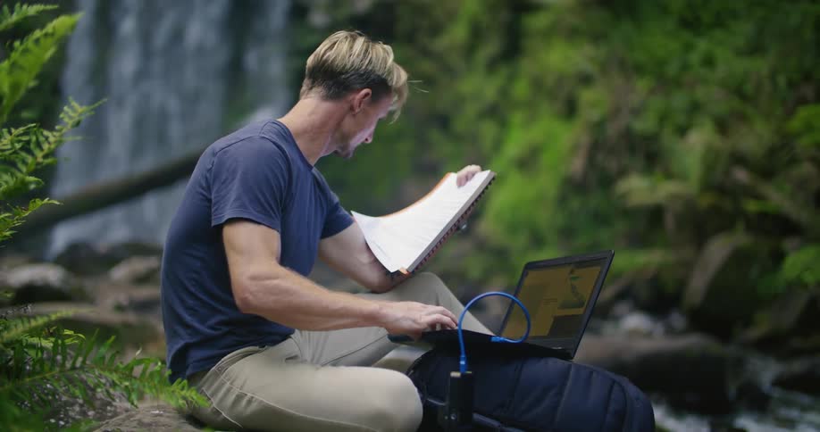 A man is sitting on a ledge near waterfalls and a stream holding papers and working on a laptop computer connected to another device
