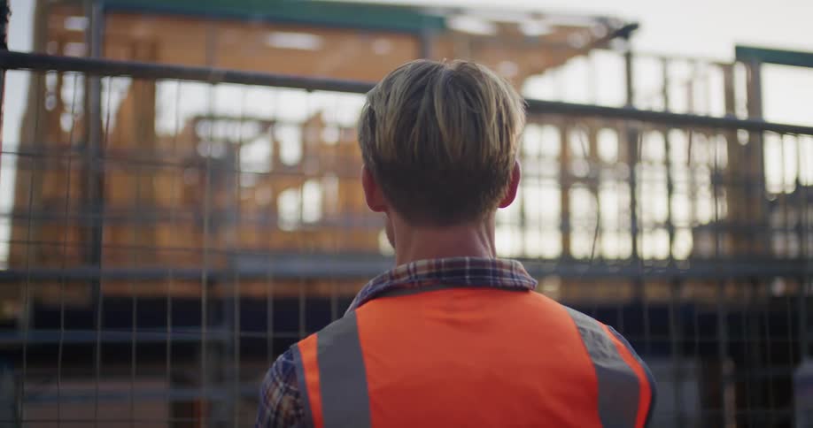 Worker stands looking through mesh fence at building under construction