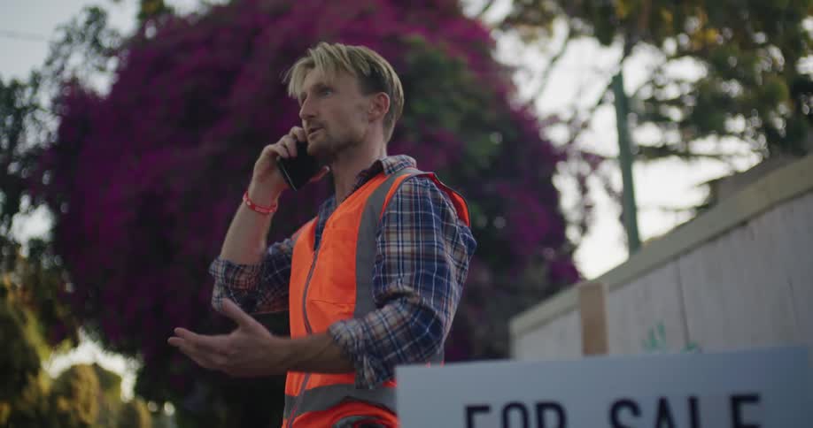 Man wearing safety vest stands near for sale sign for a foreclosed property while talking on the phone