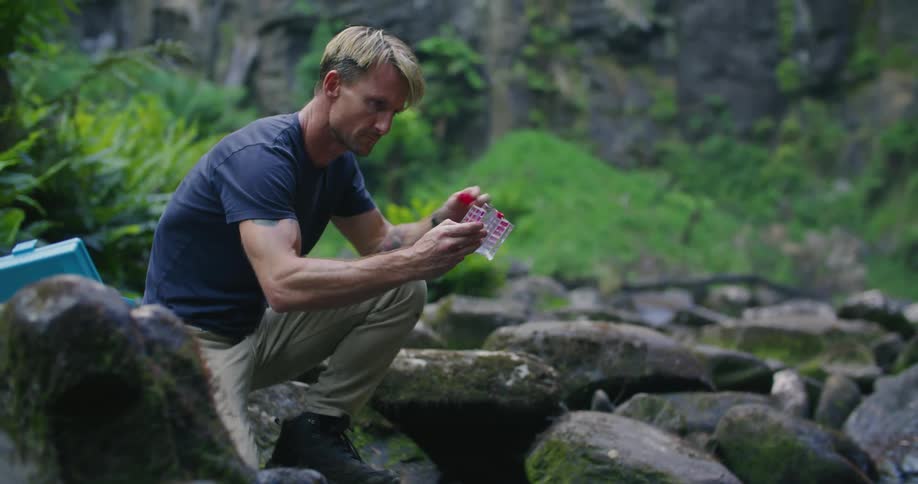 A man is kneeling on rocks at the side of a flowing stream in the woods collecting a water sample in a clear container