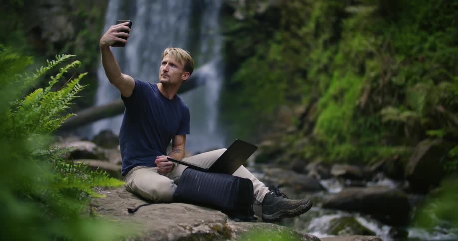 Man sits on stone on riverbank near waterfall raising device to get data before typing on laptop