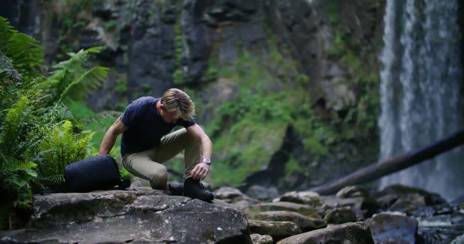 A man sits down on a large rock in front of a waterfall and opens his backpack