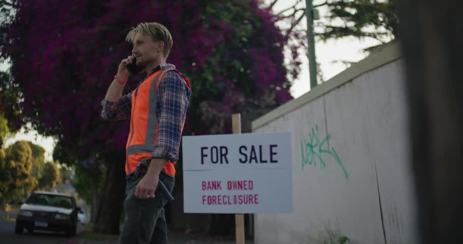 A construction worker in an orange vest making a phone call next to a posted for sale sign