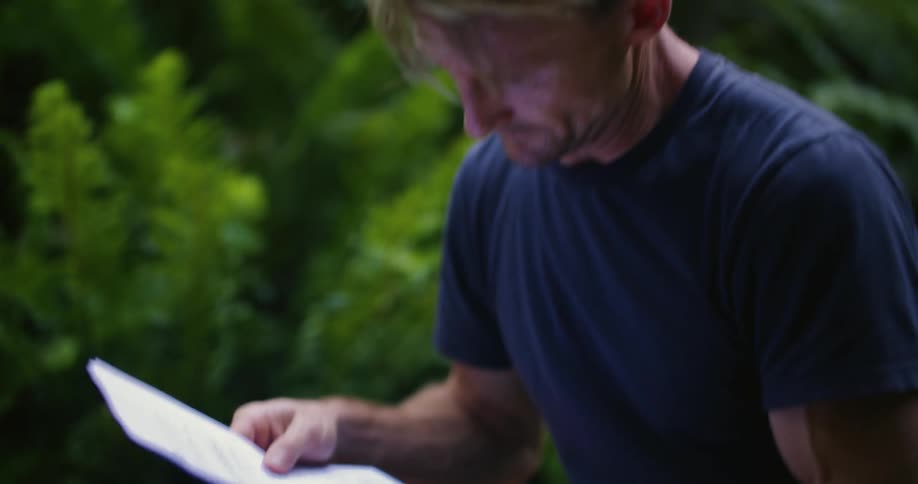Man sits outdoors reading paperwork and typing on laptop
