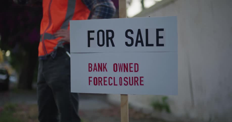 A man wearing a bright red vest is standing next to a sign in front of a house talking on his phone