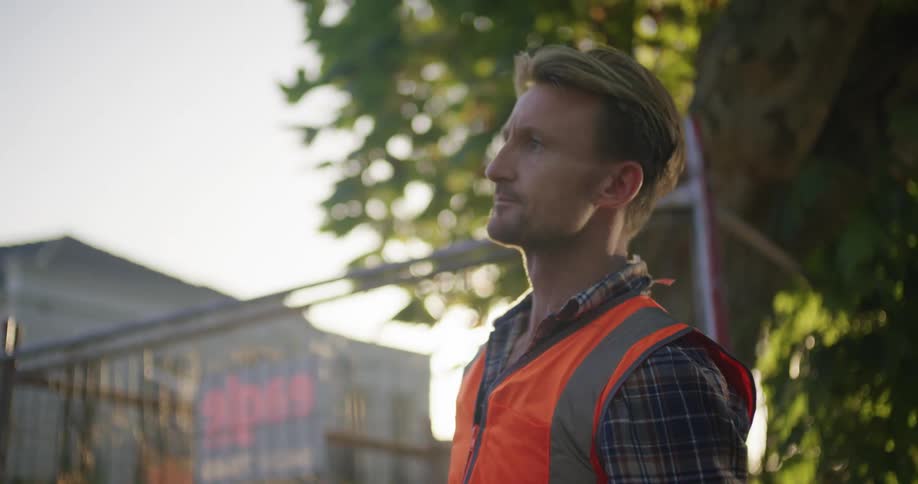 A man wearing a bright red vest is standing in front of a house and pounding a sign into the ground with a hammer