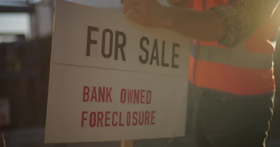 Man wearing safety vest hammers for sale sign for foreclosed home into ground