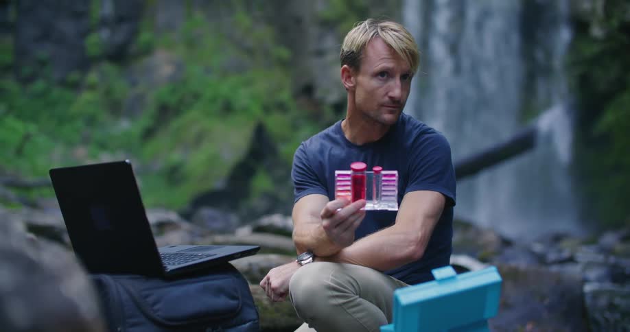 Man crouches next to laptop near waterfall and contemplates water and water testing kit