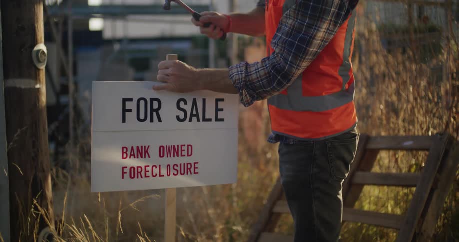 A construction worker in an orange vest hammering a for sale sign into the ground