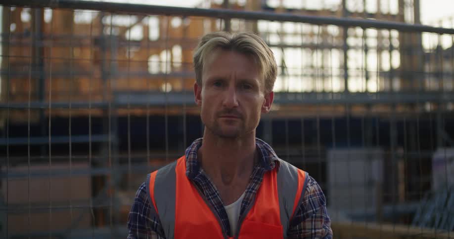 Man in plaid shirt and safety vest smiles at camera in the construction area at sunset