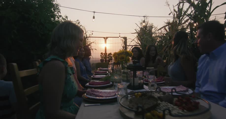 Two children are outside running on a path to the family sitting around a long dining table as sun shines brightly in the distance