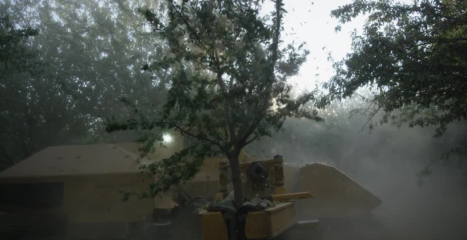 Agricultural machinery clamps padded arms onto tree trunk and shakes it causing leaves to fall on hazy day
