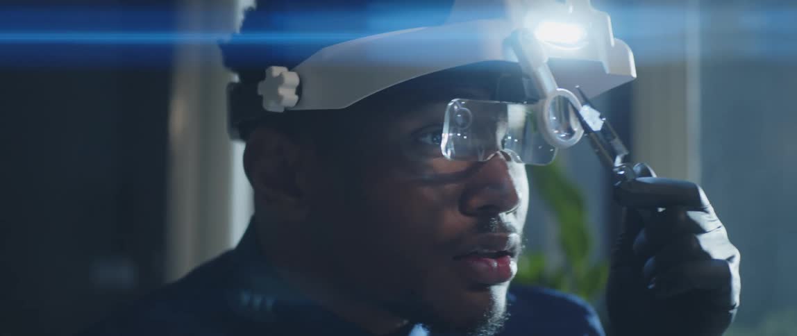 A man is holding an object with pliers as he looks at through a magnifying glass on his helmet