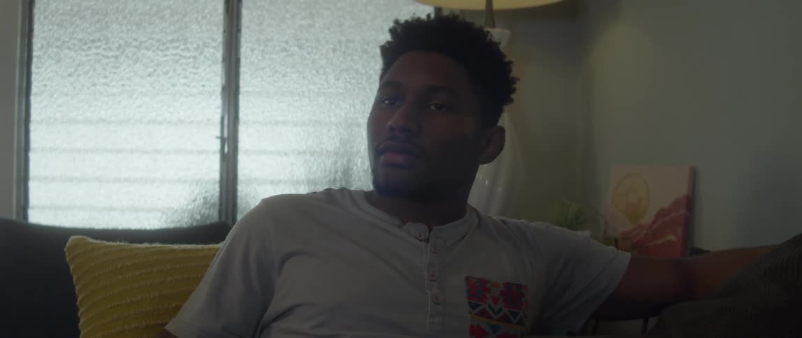 A man is sitting on a couch and he picks up his phone and looks at it then shakes his head and sets the phone down