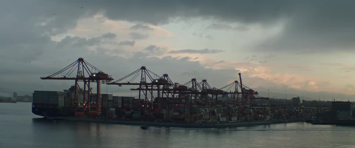 Motorboat moves past loading cranes on dock under a cloudy sky