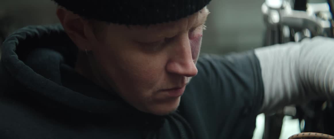 A mechanic wrenches on her motorcycle wearing hat and gray hoodie