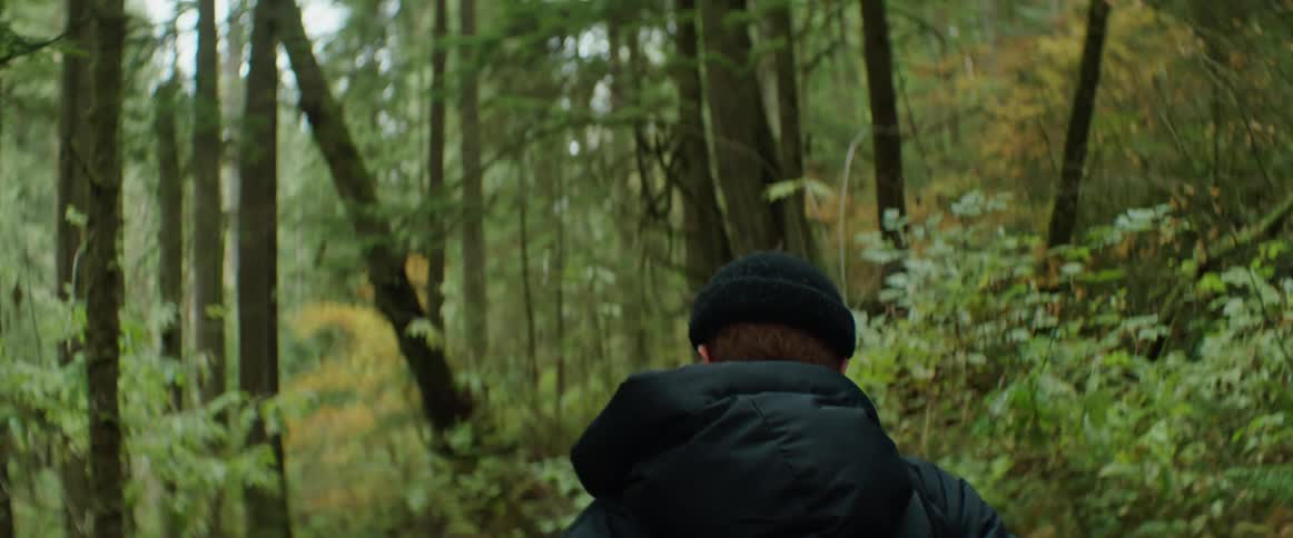 A woman wearing a black coat with a hood and a knit cap is walking through the forest