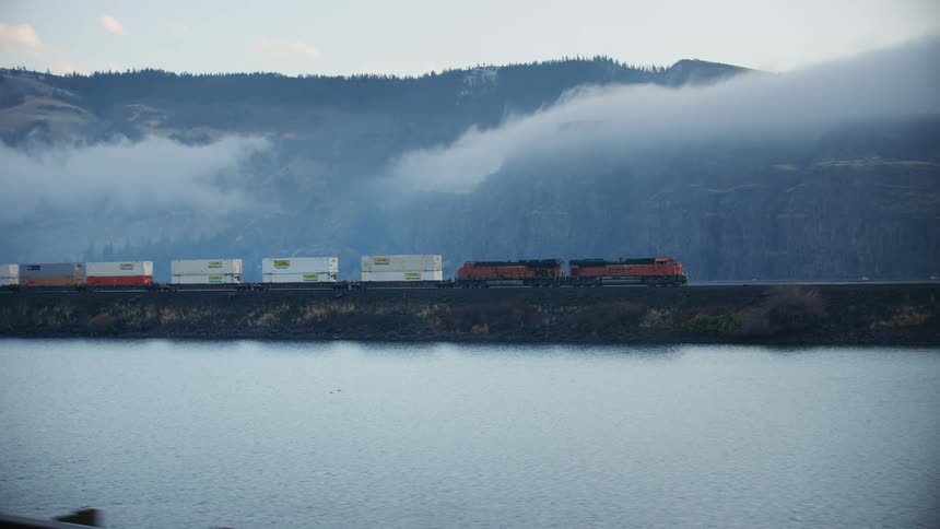 Freight train runs along railway track near the lake and mountains on a foggy and beautiful day