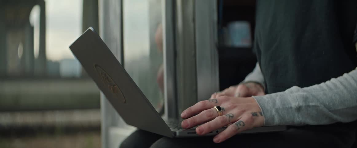 A person wearing a long sleeve shirt and is sitting on a vehicle typing on a laptop computer