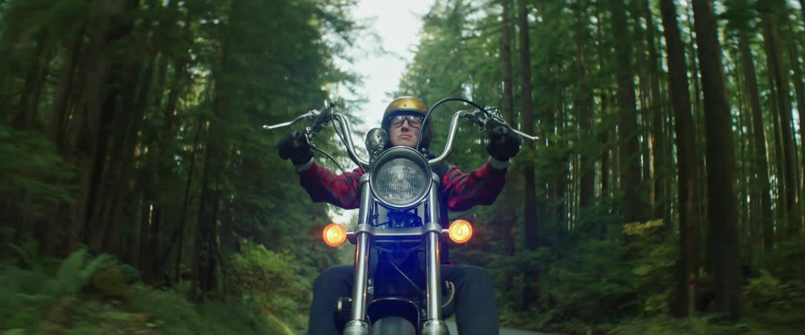 Woman with helmet red plaid shirt and pants rides a chopper on highway through forest