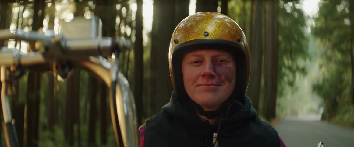 A person wearing a yellow helmet sitting on a motor bike in the middle of the road among tall trees