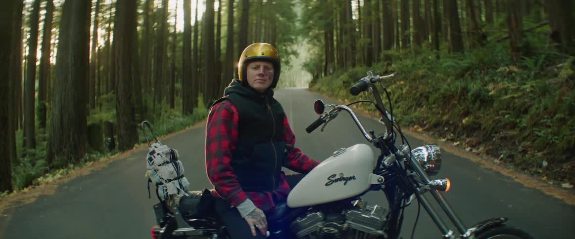 A woman wearing a gold helmet is sitting on a motorcycle parked across a forest road