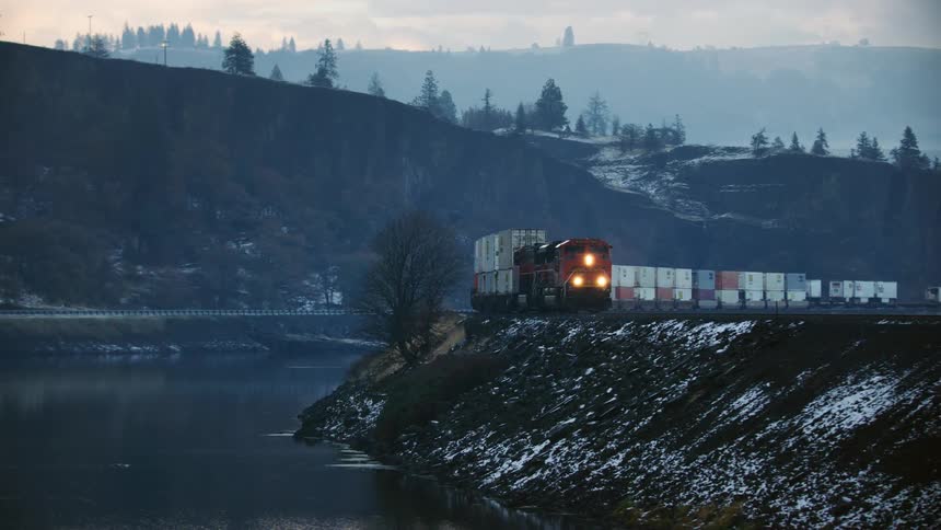 A freight train pulling cargo is going around a curve above a river near forested mountains with houses on the hillside