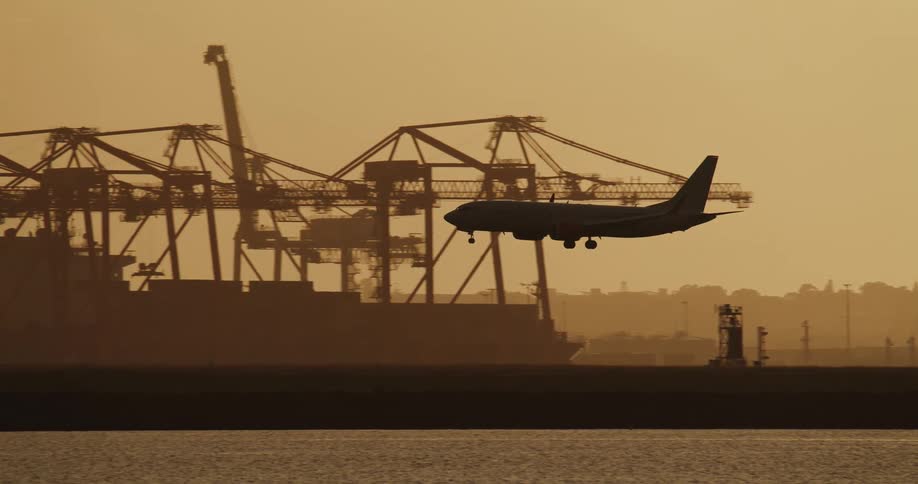 The silhouette of a passenger jet flying low with landing gear down past shipping cranes
