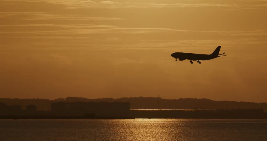 An airplane flies over the coast as the sun sets