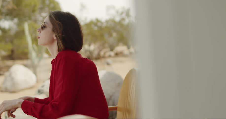Woman wearing bright red clothes and shades leans forward in chair on patio near landscaped rock garden