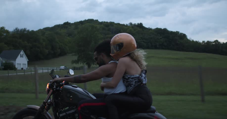 Two people are riding a motorcycle on a rural road as a woman has her arms around the mans waist as his hands are on the handlebars