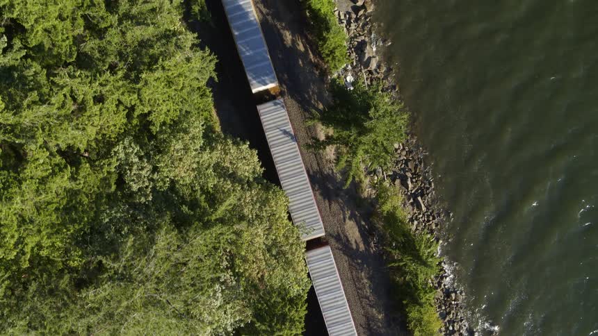 Following from above as boxcars roll down a track between trees and water