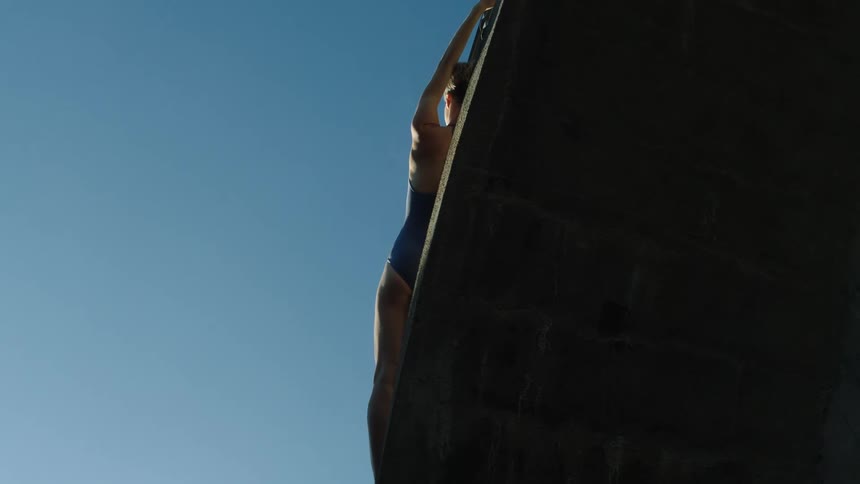 The sun shines upon a woman in a bathing suit stretched out on the edge of a ledge under a clear sky